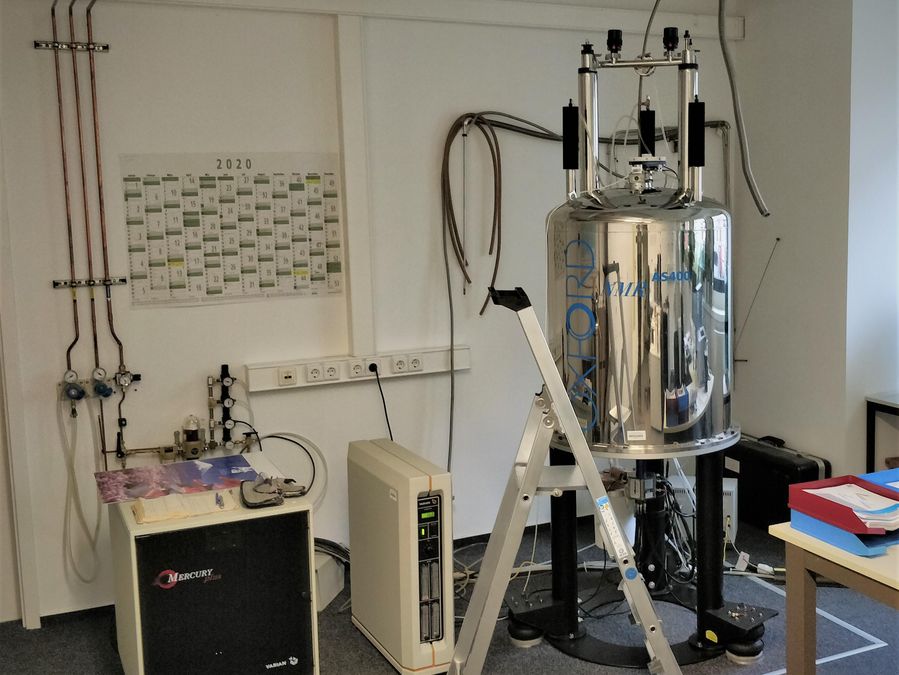 zur Vergrößerungsansicht des Bildes: NMR-Spektrometer mit Chromglänzender Oberfläche und angeschlossener Peripherie, im Vordergrund eine Leiter