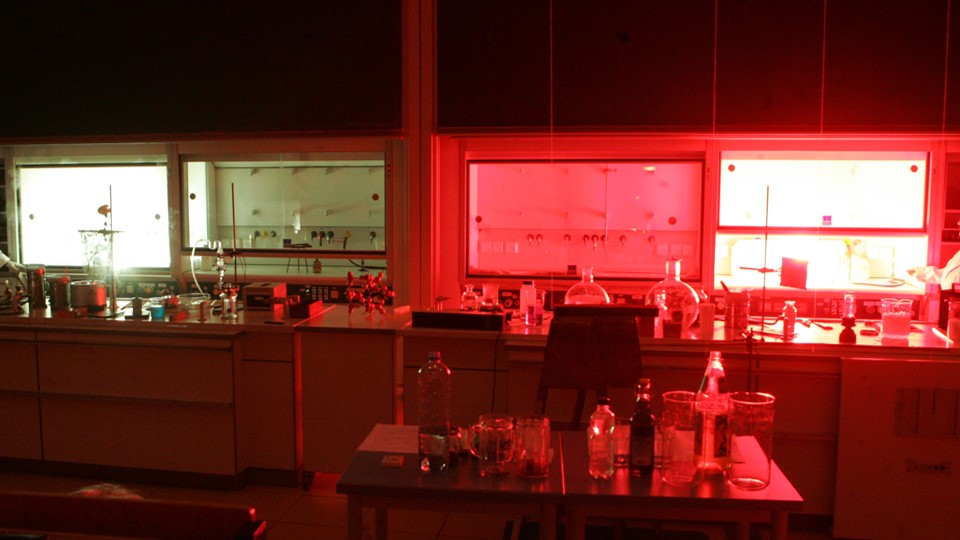 zur Vergrößerungsansicht des Bildes: Abgedunkelter Chemiehörsaal, Glasgeräte auf dem Pult, Rot und Grün leuchtende Abzüge