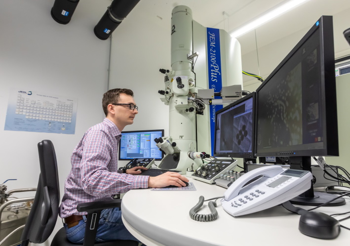 Person sitzt an einem Arbeitsplatz mit zwei Bildschirmen und Bedieneinheiten, im Hintergrund ein Transmissionselektronenmikroskop